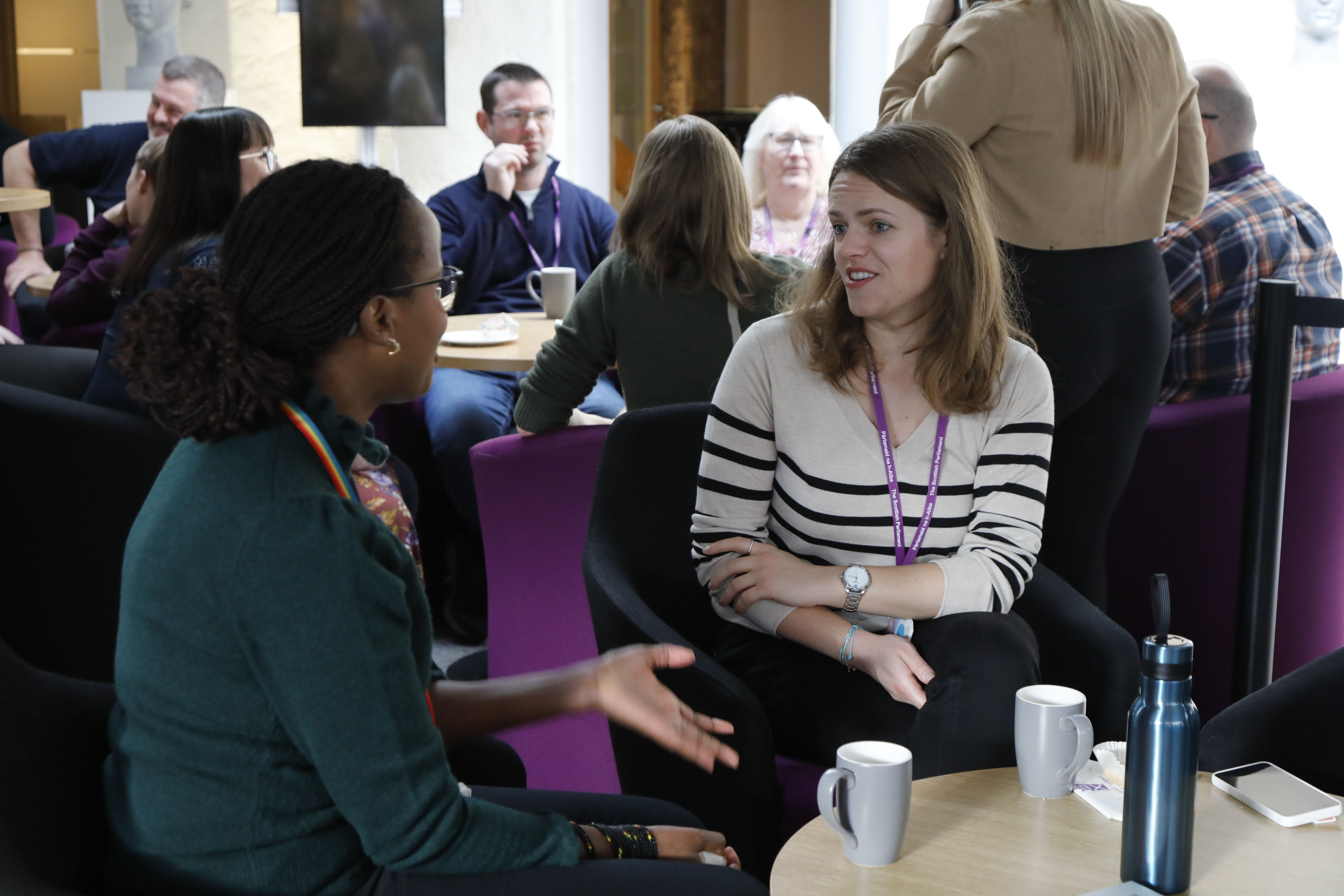 Two women chatting at a coffee table. There are other members of staff talking in the background.