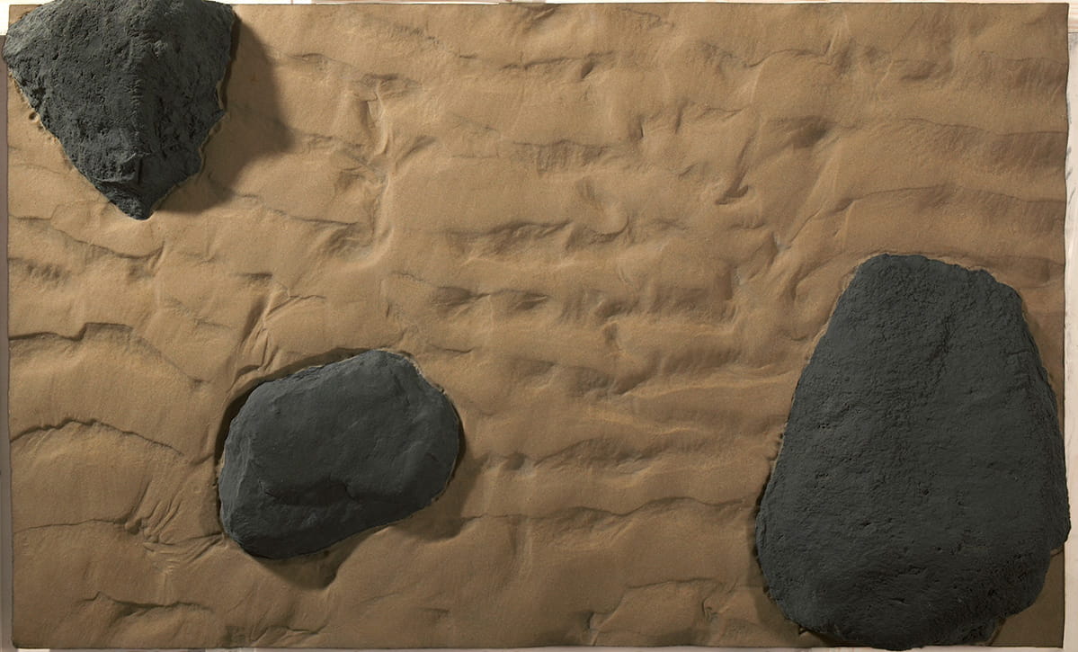 Fibreglass cast of a segment of sand and rocks, painted in realistic colours. Three black rocks protrude from rippled golden colour sand. 