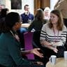 Two women sitting down and chatting. Other staff members are having a meeting behind them.